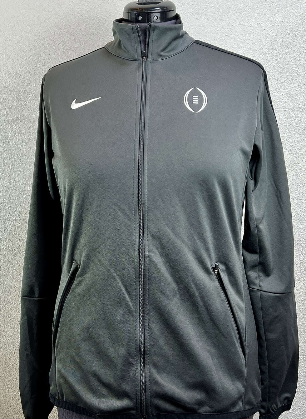 A front view of a Nike full-zip jacket in grey, featuring the Nike logo on the right chest and a College Football Playoff emblem on the left. The jacket has a high collar, zippered pockets, and is displayed on a mannequin against a neutral background.