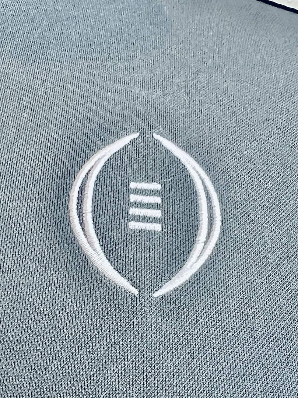 A close-up of the College Football Playoff emblem on a grey jacket. The emblem is a stylized football design above three horizontal lines, suggesting a stadium, embroidered in white thread.