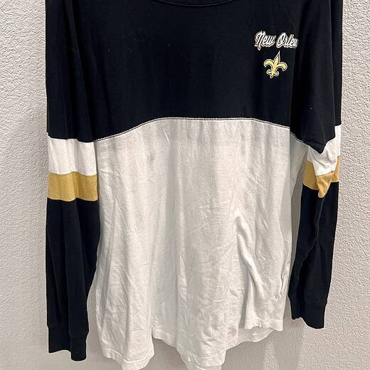 Front view of a New Orleans Saints long sleeve tee, showcasing bold ‘SAINTS’ lettering in white with gold trim on a black background.