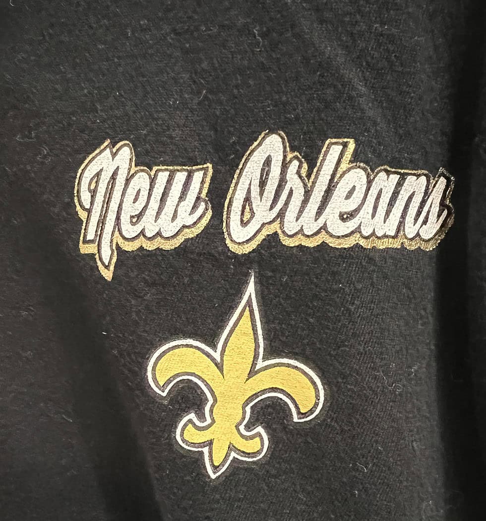 Close-up of the upper-left chest area with the embroidered ‘New Orleans’ script and Saints fleur-de-lis logo in gold.