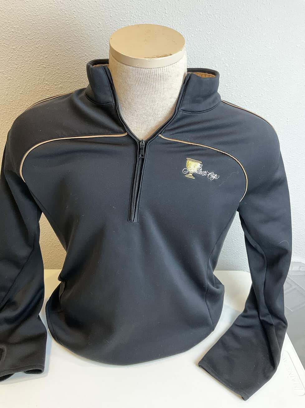 Black quarter-zip pullover with gold trim detailing and the Presidents Cup logo embroidered on the upper left chest, designed with a high collar.