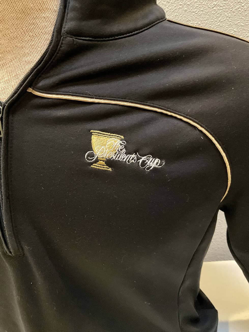 Close-up of the Presidents Cup logo embroidered on a black pullover, detailed with a golf trophy and the event's name, set against a black fabric with a gold trim accent.
