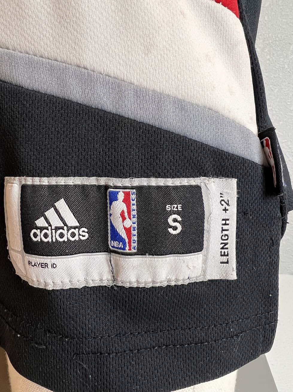 Close-up of the size tag on a black Adidas Portland Trail Blazers NBA jersey, indicating 'SIZE S' (Small), 'LENGTH +2'. The tag also includes the Adidas logo, NBA logo, and a 'PLAYER ID' field.