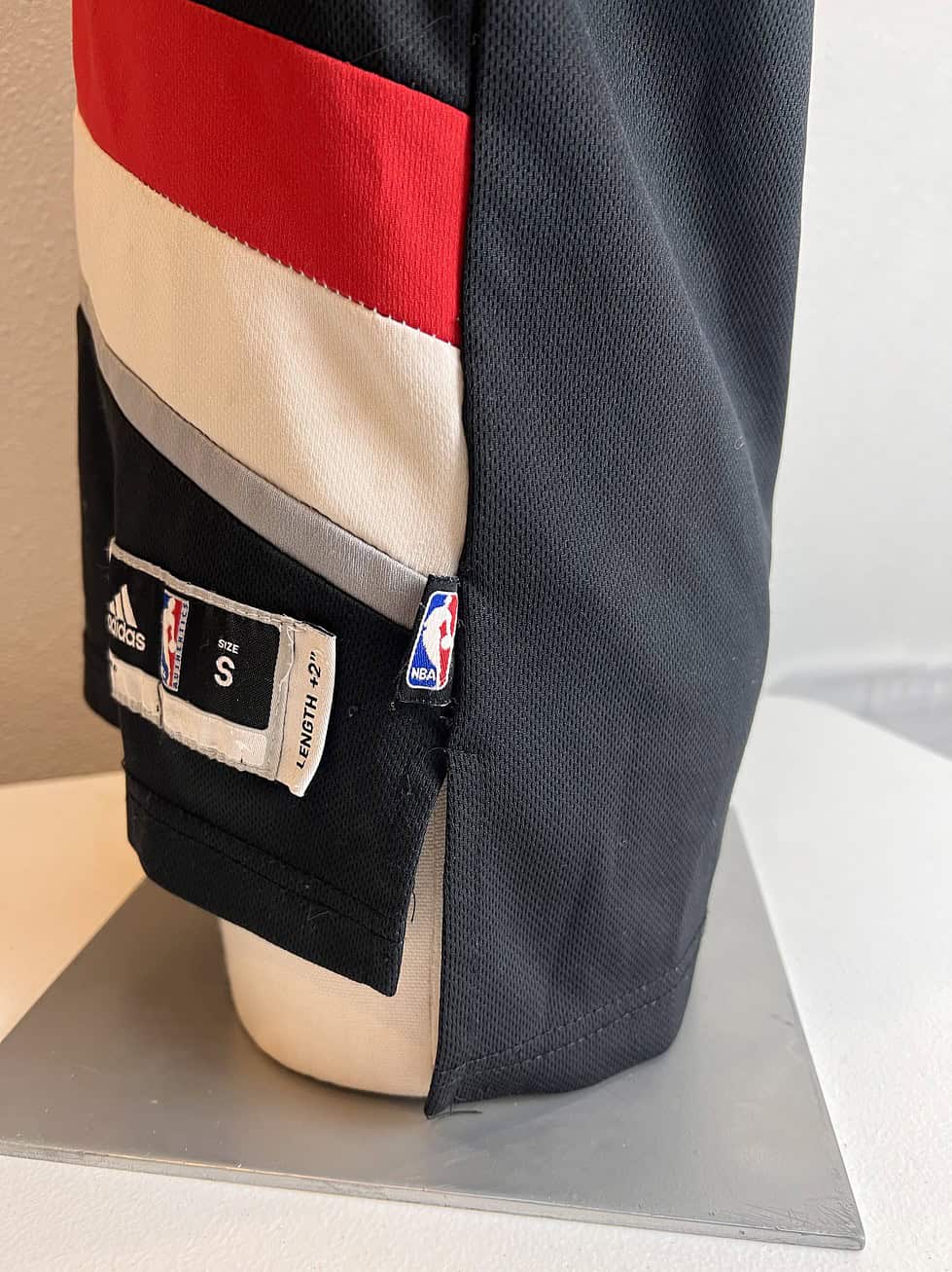 Close-up of the size tag on a black Adidas Portland Trail Blazers NBA jersey, indicating 'SIZE S' (Small), 'LENGTH +2'. The tag also includes the Adidas logo, NBA logo, and a 'PLAYER ID' field.