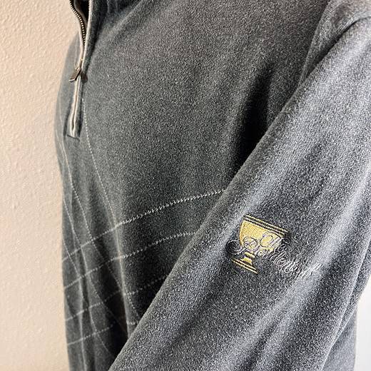 Close-up of the sleeve of a charcoal grey sweater, displaying an embroidered Presidents Cup trophy logo, signifying the prestigious golf event.