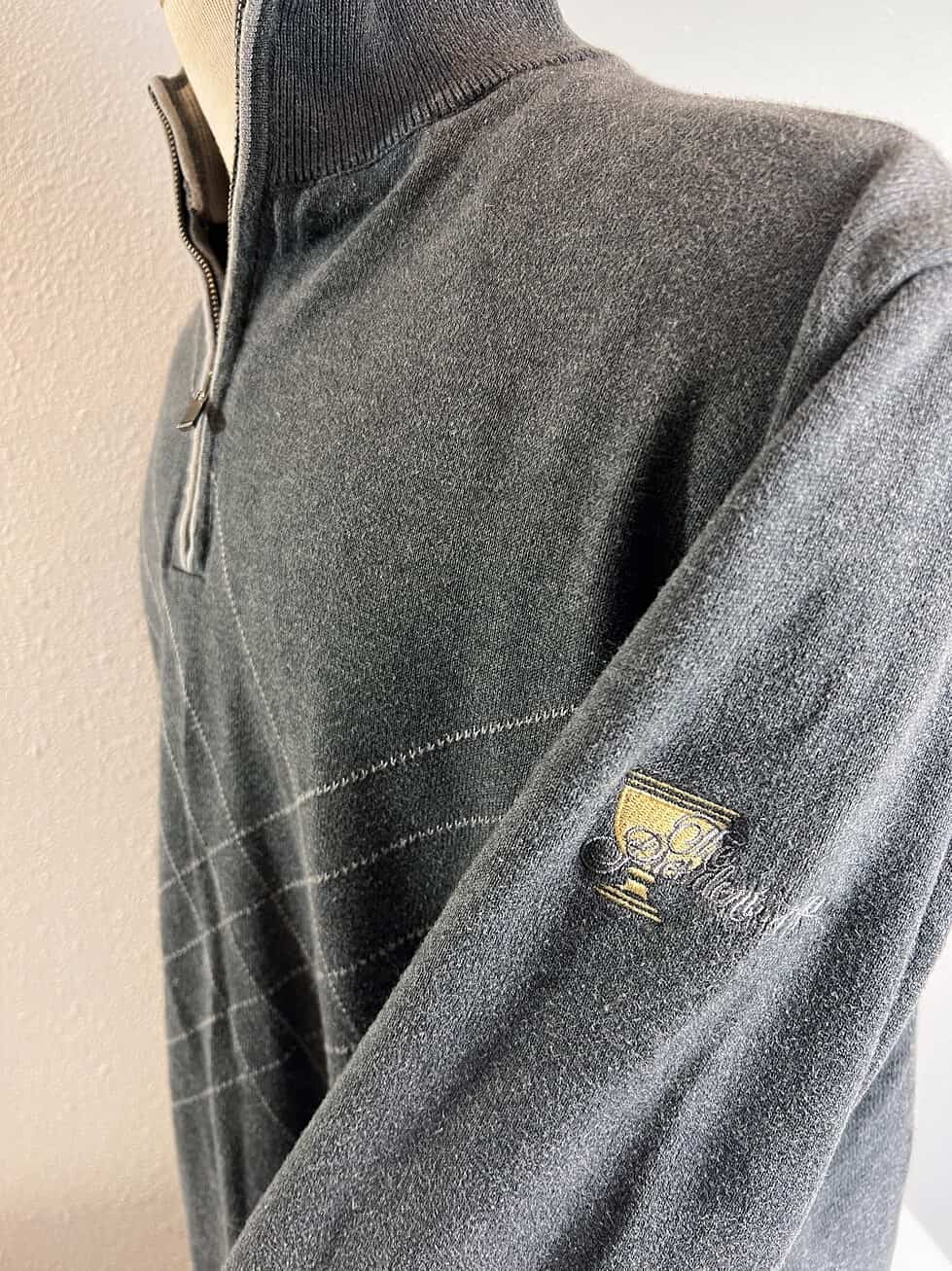 Close-up of the sleeve of a charcoal grey sweater, displaying an embroidered Presidents Cup trophy logo, signifying the prestigious golf event.