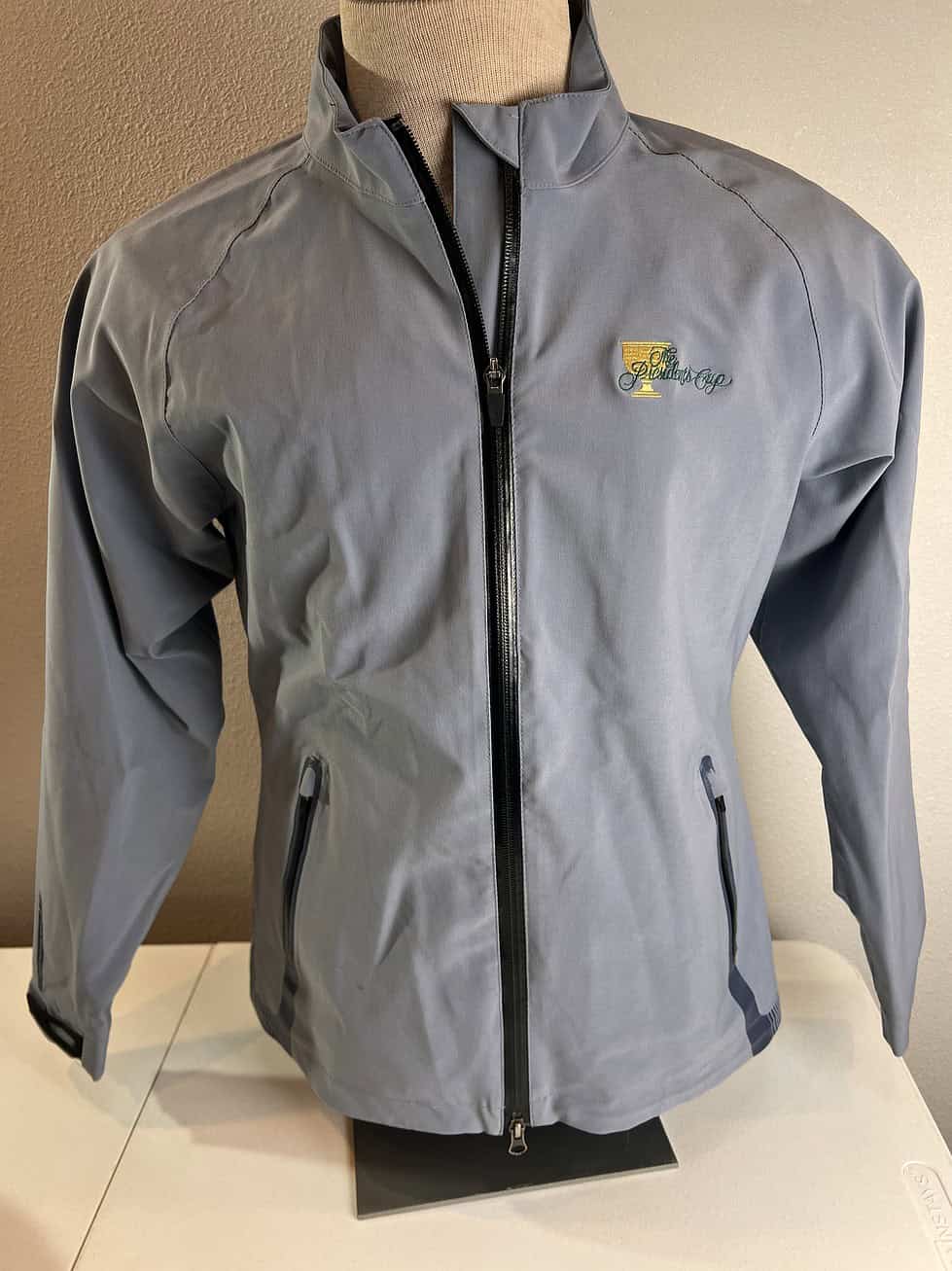 Light grey Sun Mountain rain jacket with a full zipper front, zippered pockets, and the 2013 Presidents Cup logo embroidered on the chest.