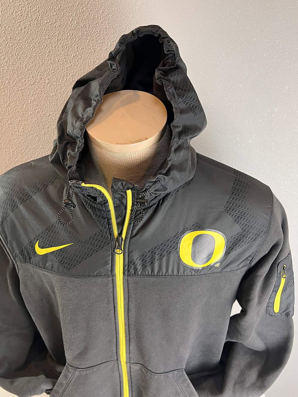 Charcoal grey Nike hoodie with a bright yellow zip and accents, featuring the University of Oregon 'O' emblem on the right chest area and a Nike swoosh on the left. The jacket has a textured upper panel, a hood, and front pockets.