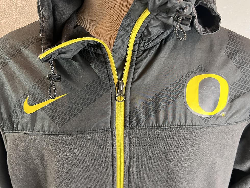 Close-up of the upper part of a grey Nike hoodie, highlighting the yellow Nike swoosh logo on the left chest and the University of Oregon 'O' emblem on the right, with a detailed textured pattern across the shoulders and hood.