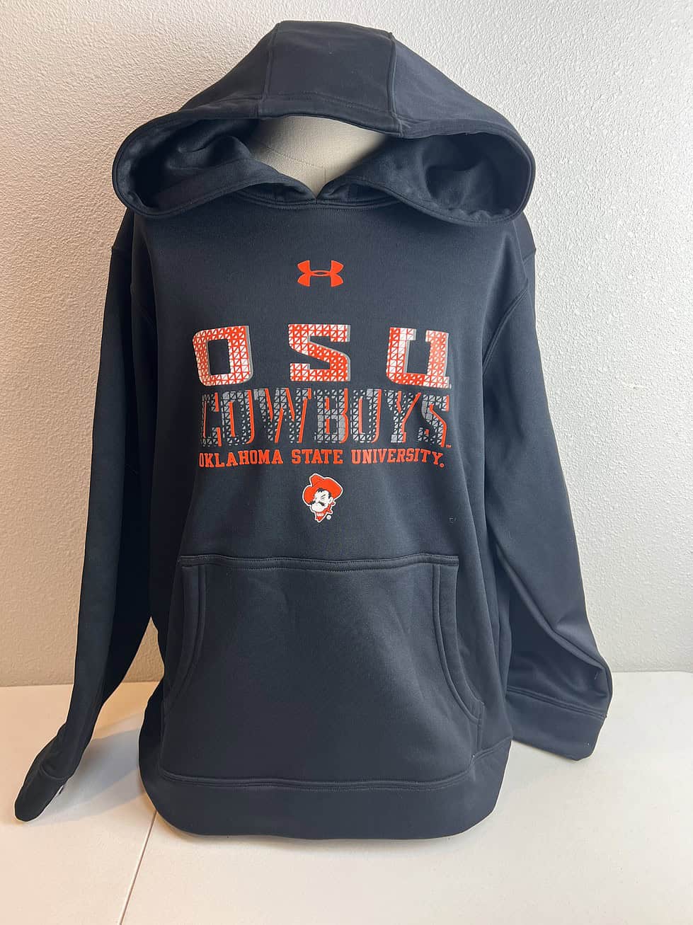 Youth-sized Under Armour hoodie in black, featuring 'OSU COWBOYS' lettering and Oklahoma State University mascot logo in orange, with a front pouch pocket.