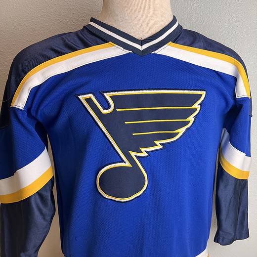 Front view of a St. Louis Blues hockey jersey in blue with the team’s logo, yellow and white stripes on the sleeves, and a Starter brand logo on the hem.