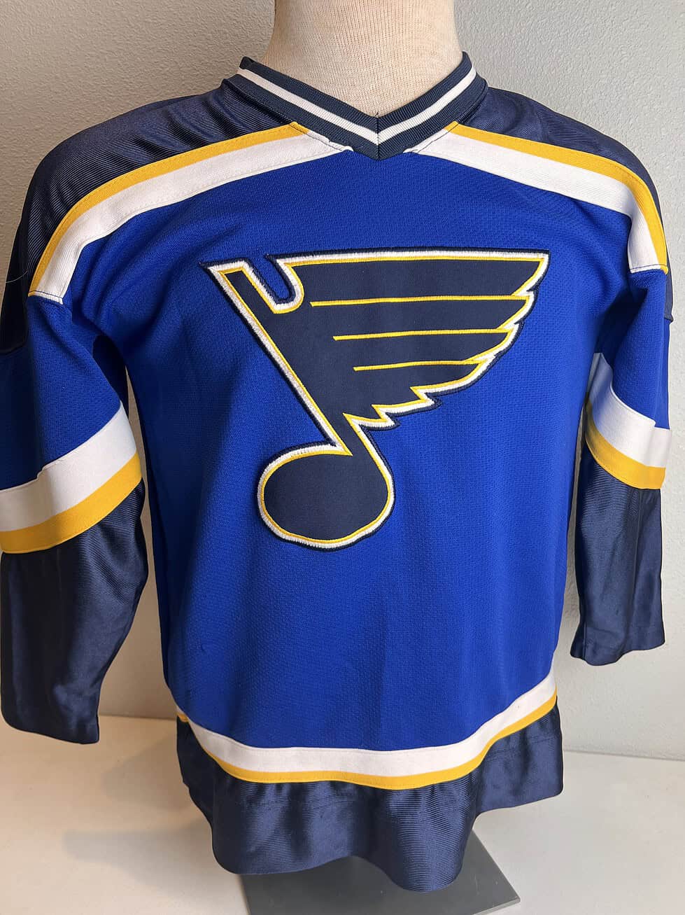 Front view of a St. Louis Blues hockey jersey in blue with the team’s logo, yellow and white stripes on the sleeves, and a Starter brand logo on the hem.