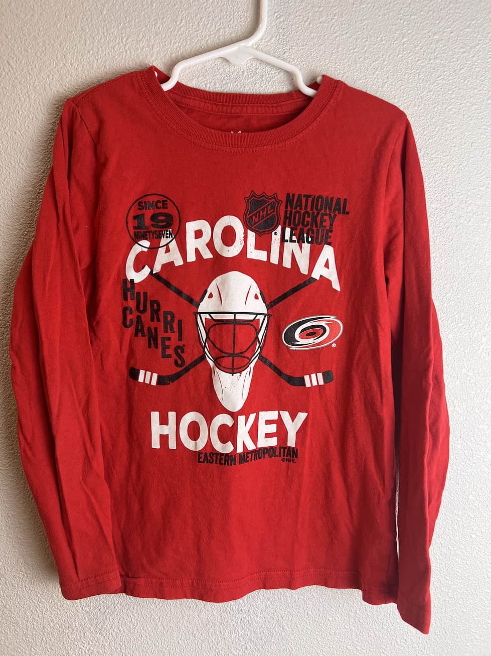 Full view of the front of a red Carolina Hurricanes youth long sleeve shirt with hockey and team-themed graphics.