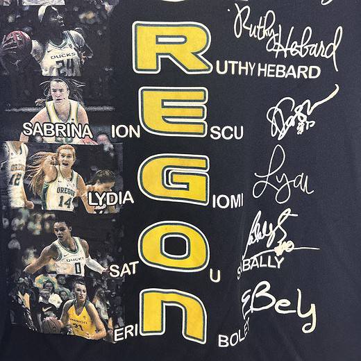 Close-up of a black Oregon Ducks basketball fan t-shirt with a yellow and white graphic design. The front features printed signatures of the 2020 team members around a vibrant 'OREGON' text and images of players in action.