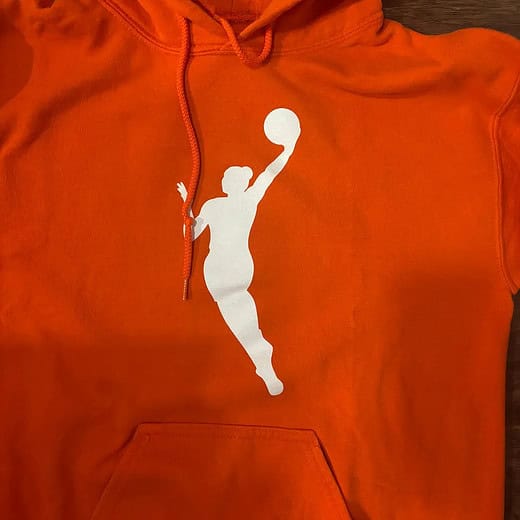 Close-up of a bright orange hooded sweatshirt featuring a white silhouette of a basketball player in mid-shot, symbolizing dynamic on-court action