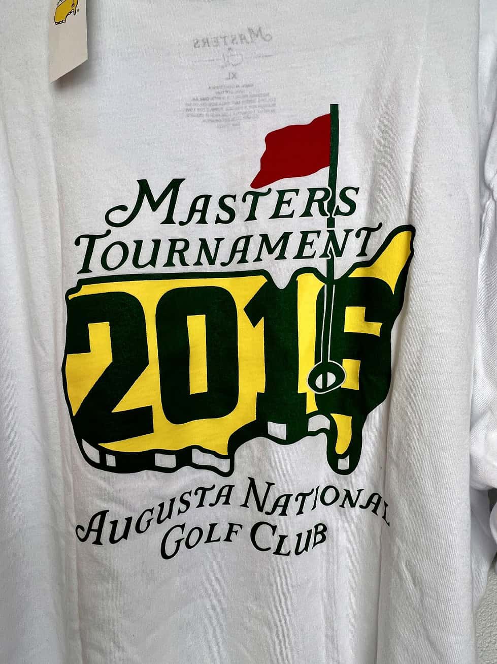 Close up view of Masters Tournament 2016 t-shirt in white. Augusta National logo in yellow with "2016" across the design. The words "Augusta National Golf Club" in brand script are underneath the logo