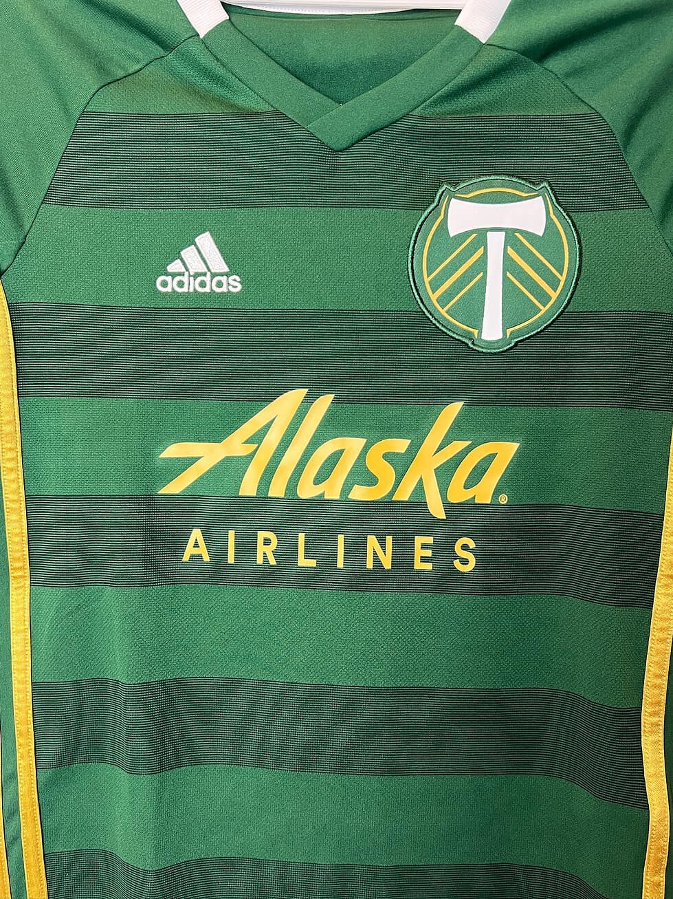 Close-up of the Portland Timbers crest and Adidas logo on a green youth jersey with gold stripes.