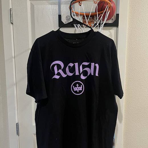 Front view of a black Stadium Essentials T-shirt with light purple "Reign" graphic and crown logo, commemorating Megan Rapinoe's career.