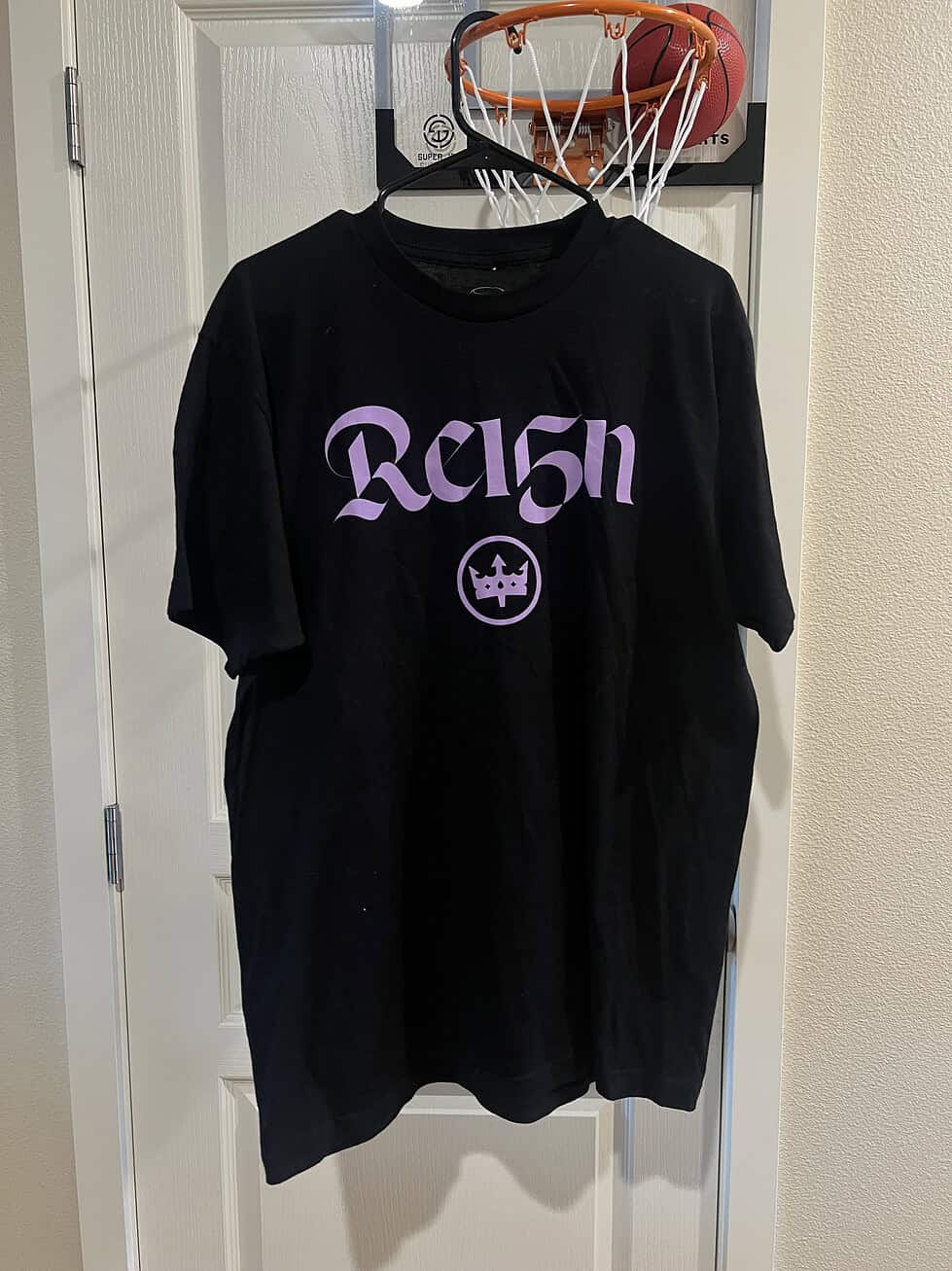Front view of a black Stadium Essentials T-shirt with light purple "Reign" graphic and crown logo, commemorating Megan Rapinoe's career.