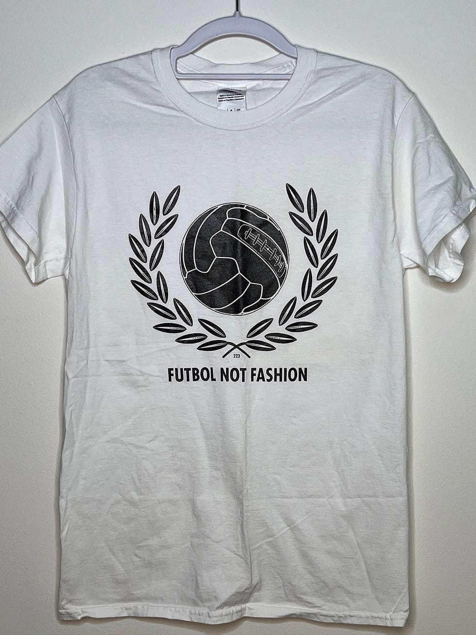 Front of t-shirt. Text says Futbol not Fashion. Image is a white and grey ball with branches on either side