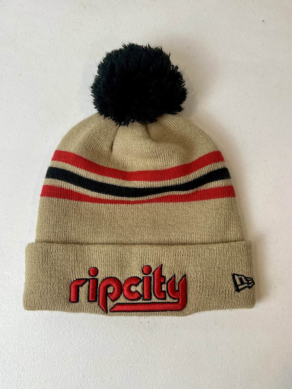 Front view of tan Rip City New Era beanie with red "ripcity" embroidery and black pom.