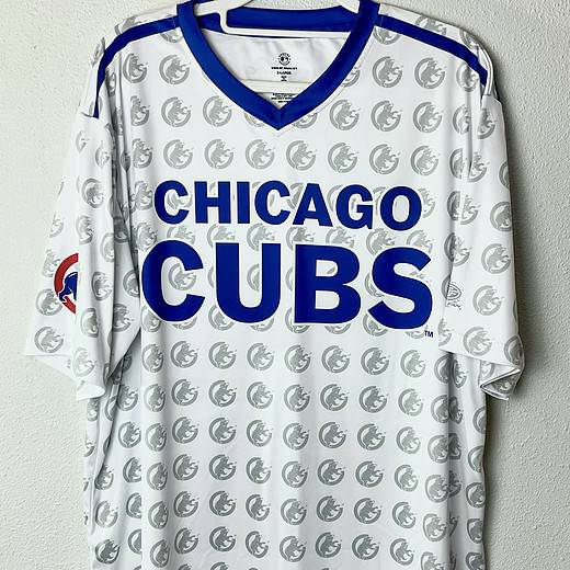 White V-neck tee with bold blue “CHICAGO CUBS” lettering across the chest and an all-over Cubs bear head watermark print. Blue shoulder accents and Cubs logo patch on the left sleeve.
