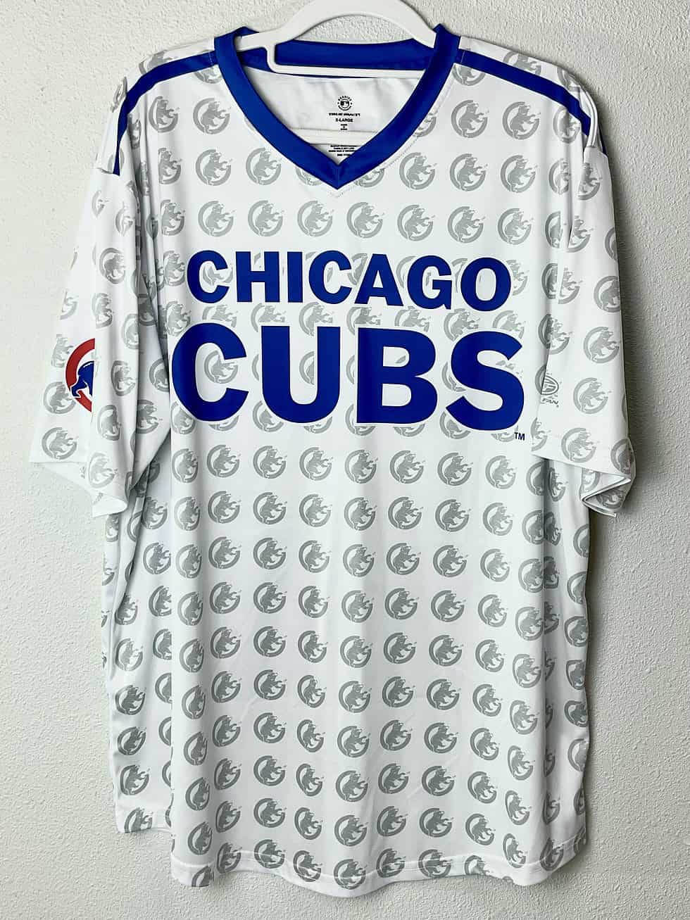 White V-neck tee with bold blue “CHICAGO CUBS” lettering across the chest and an all-over Cubs bear head watermark print. Blue shoulder accents and Cubs logo patch on the left sleeve.