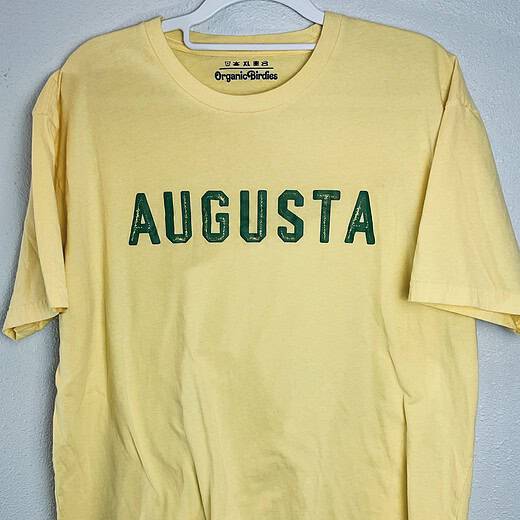 A short-sleeve yellow t-shirt with green “Augusta” lettering, displayed on a hanger against a neutral background.
