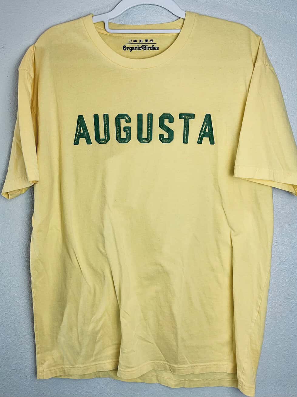 A short-sleeve yellow t-shirt with green “Augusta” lettering, displayed on a hanger against a neutral background.