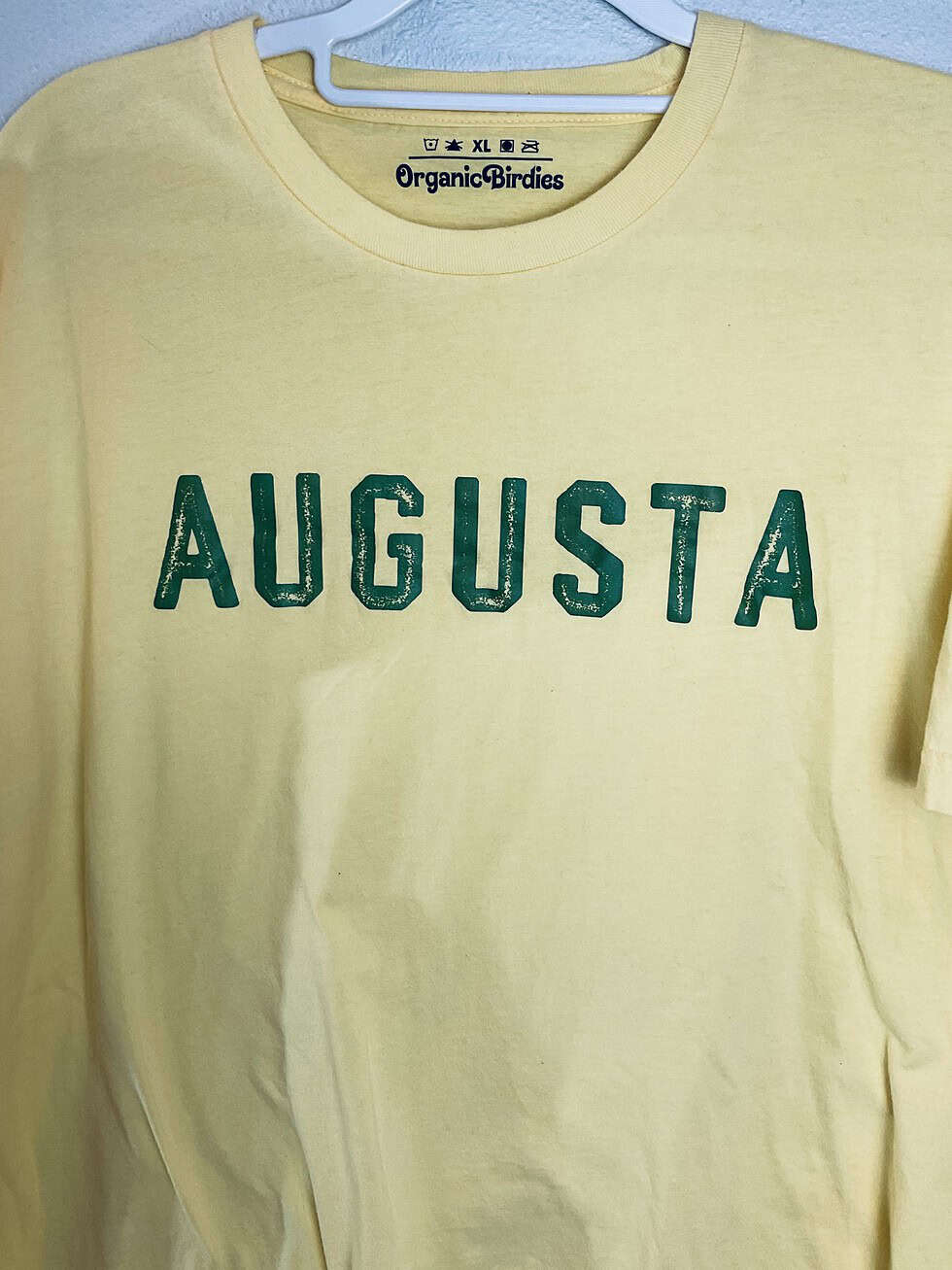 A soft yellow t-shirt featuring the word “Augusta” in distressed green block letters across the chest, made by Organic Birdies.