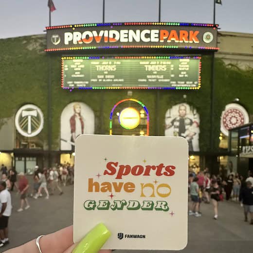 hand holds a beige sticker with the words "Sports have no gender" and FanWagn logo, positioned in front of Providence Park entrance at dusk.